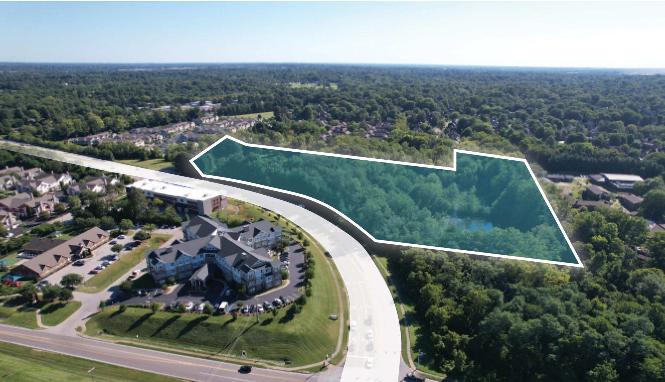 Aerial view of a lush green wooded area outlined in white, adjacent to a curved road and suburban neighborhood with houses and apartment complexes.